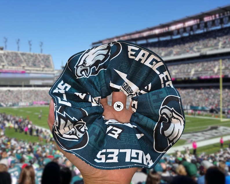 Puede incluir: Una scrunchie verde azulado y blanca con el logotipo y el nombre del equipo de los Philadelphia Eagles. La scrunchie se sostiene frente a un estadio de f&uacute;tbol.