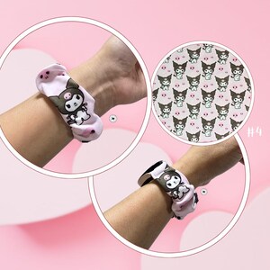 Op de afbeelding: Roze scrunchie met een herhaald patroon van een stripfiguur met zwarte oren en een wit lichaam. De scrunchie is om een pols gewikkeld. Een cirkelvormige afbeelding toont het patroon van dichtbij.