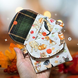 May include: A multi-compartment wallet with a green plaid flap and a dog-themed print. The print features various dog breeds, pumpkins, and fall leaves. The wallet has orange snap closures and is held open.