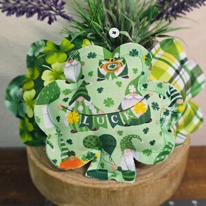 Puede incluir: Un adorno de tela en forma de trébol verde con un borde de tela a cuadros verde, blanco y amarillo. El trébol está decorado con gnomos, tréboles y la palabra "LUCK" en una pancarta.