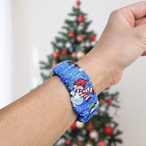 May include: A blue scrunchie with a repeating pattern of snowmen, snowflakes, and Christmas trees. The scrunchie is wrapped around a wrist, with a Christmas tree in the background.