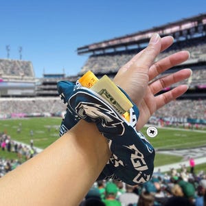 Puede incluir: Una scrunchie azul marino y blanca con cremallera, con el logotipo de los Philadelphia Eagles. La scrunchie est&aacute; envuelta alrededor de una mu&ntilde;eca y contiene efectivo y una tarjeta de cr&eacute;dito. El fondo muestra un estadio de f&uacute;tbol.