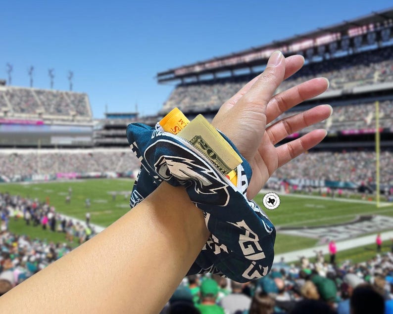 Puede incluir: Una scrunchie azul marino y blanca con cremallera, con el logotipo de los Philadelphia Eagles. La scrunchie est&aacute; envuelta alrededor de una mu&ntilde;eca y contiene efectivo y una tarjeta de cr&eacute;dito. El fondo muestra un estadio de f&uacute;tbol.