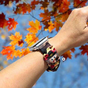 Op de afbeelding: Een zilveren smartwatch met een patroonband met een cartoon kalkoen ontwerp. De band is bruin, zwart en bruin met een kalkoen met een pelgrimshoed. De achtergrond toont herfstbladeren.