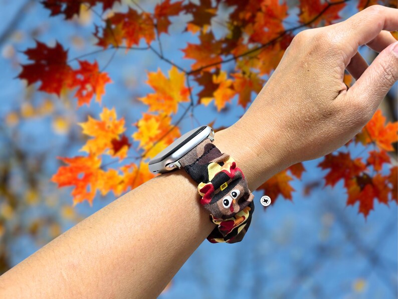Op de afbeelding: Een zilveren smartwatch met een patroonband met een cartoon kalkoen ontwerp. De band is bruin, zwart en bruin met een kalkoen met een pelgrimshoed. De achtergrond toont herfstbladeren.