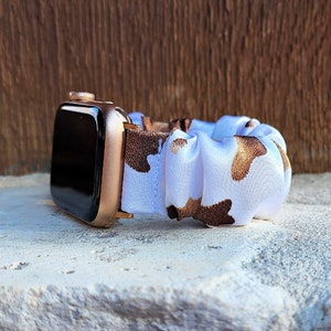 Op de afbeelding: Een smartwatch met een metallic ros&eacute;gouden behuizing en een zwart scherm, bevestigd aan een stoffen scrunchie horlogebandje met een witte achtergrond en een bruin met beige koeienvlekkenpatroon in een geplooid ontwerp.