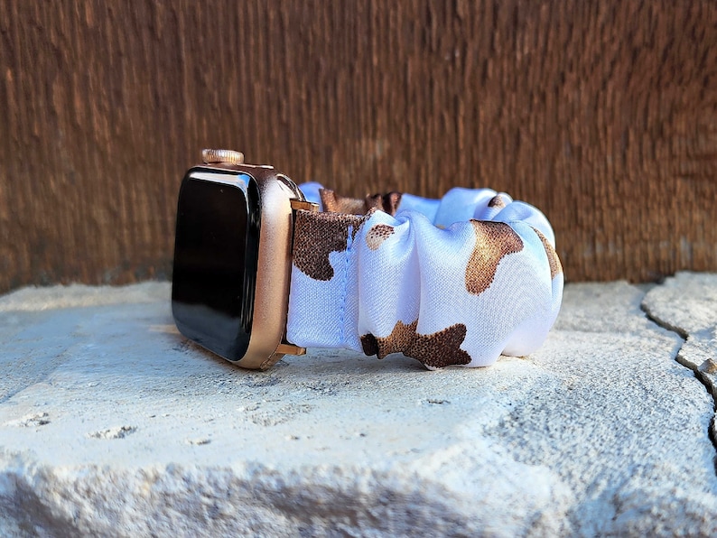 Op de afbeelding: Een smartwatch met een metallic ros&eacute;gouden behuizing en een zwart scherm, bevestigd aan een stoffen scrunchie horlogebandje met een witte achtergrond en een bruin met beige koeienvlekkenpatroon in een geplooid ontwerp.