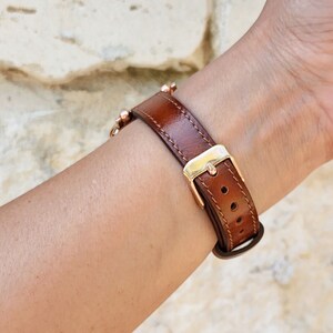 May include: A brown leather watch band with a gold-colored buckle and a series of small, gold-colored beads. The band is secured with a buckle and has several holes for adjustment. The watch band is designed to be worn on the wrist.