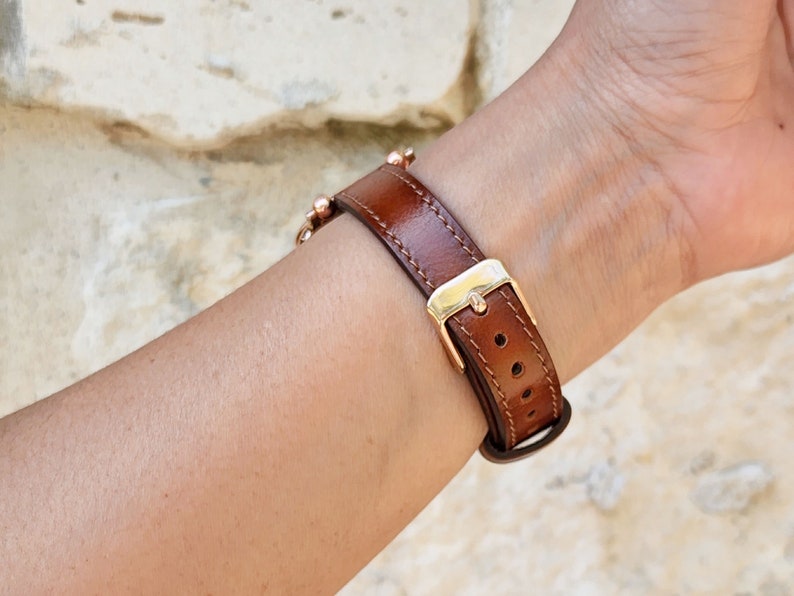 May include: A brown leather watch band with a gold-colored buckle and a series of small, gold-colored beads. The band is secured with a buckle and has several holes for adjustment. The watch band is designed to be worn on the wrist.