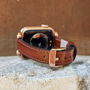 May include: A brown leather watch band with a gold-colored buckle and stitching. The watch band is attached to a gold-colored smartwatch. The watch band is resting on a stone surface.