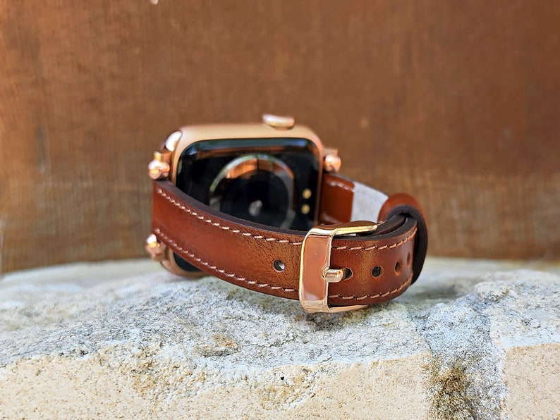 May include: A brown leather watch band with a gold-colored buckle and stitching. The watch band is attached to a gold-colored smartwatch. The watch band is resting on a stone surface.