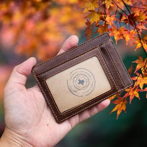 May include: A brown leather cardholder with a clear ID window. The cardholder has a stamped design inside the window. The background features autumn foliage with orange and yellow leaves.
