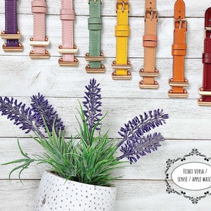 May include: A selection of watch straps in a range of colours: purple, pink, green, yellow, tan, orange, and burgundy. Each strap has a gold-coloured buckle and is set against a white wooden backdrop. A potted lavender plant is in the foreground.