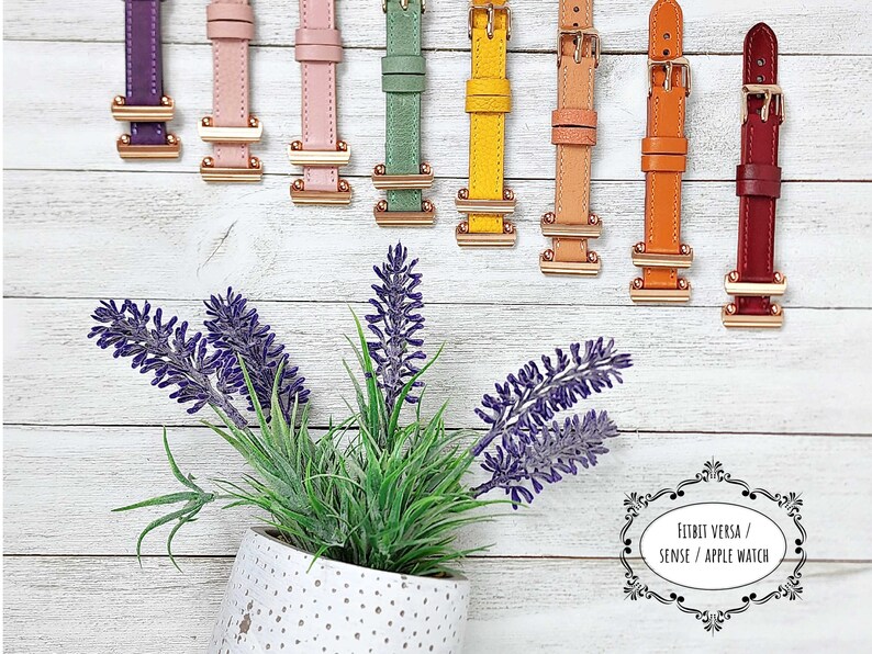 May include: A selection of watch straps in a range of colours: purple, pink, green, yellow, tan, orange, and burgundy. Each strap has a gold-coloured buckle and is set against a white wooden backdrop. A potted lavender plant is in the foreground.