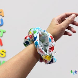 Puede incluir: Un brazalete blanco con cierre de cremallera y un estampado de huellas de manos de colores. El brazalete se lleva en la mu&ntilde;eca.