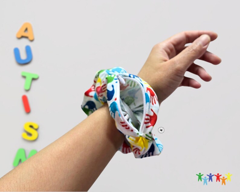 Puede incluir: Un brazalete blanco con cierre de cremallera y un estampado de huellas de manos de colores. El brazalete se lleva en la mu&ntilde;eca.