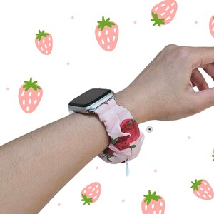Peut inclure: Bracelet de montre Apple en tissu rose avec un motif de fraises rouges, port&eacute; au poignet. Le bracelet pr&eacute;sente un motif de fraises r&eacute;p&eacute;titif. Le fond est blanc avec des illustrations de fraises.