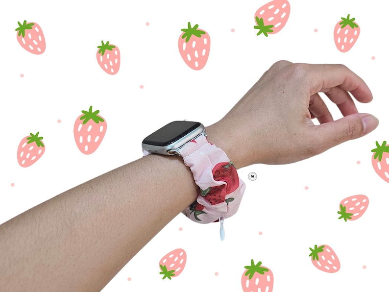 Peut inclure: Bracelet de montre Apple en tissu rose avec un motif de fraises rouges, port&eacute; au poignet. Le bracelet pr&eacute;sente un motif de fraises r&eacute;p&eacute;titif. Le fond est blanc avec des illustrations de fraises.