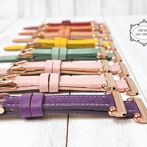 May include: A selection of colourful leather watch straps in shades like purple, pink, green, and yellow. Each strap has a rose gold buckle and connector. The straps are displayed on a white wooden surface.