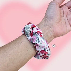 Peut inclure: Un chouchou floral avec un fond blanc et des fleurs rouges et roses est port&eacute; au poignet. Le chouchou est en tissu et a un design fronc&eacute;. Le fond est un d&eacute;grad&eacute; de rose.