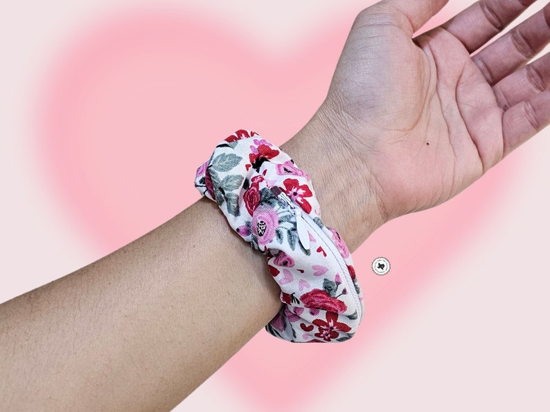 Peut inclure: Un chouchou floral avec un fond blanc et des fleurs rouges et roses est port&eacute; au poignet. Le chouchou est en tissu et a un design fronc&eacute;. Le fond est un d&eacute;grad&eacute; de rose.