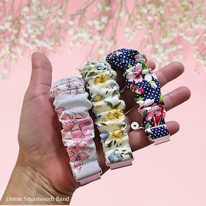 Peut inclure: Trois bracelets de montre connectée de 18 mm, chacun avec un motif floral, tenus dans une main. L'un est blanc avec des fleurs roses, un autre est jaune avec des fleurs bleues, et le troisième est bleu marine avec des fleurs rouges et des pois blancs.