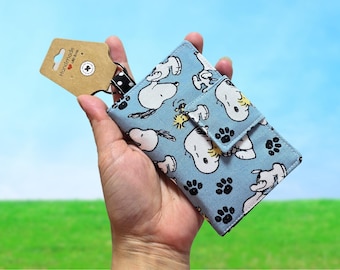 May include: A light blue wallet featuring a repeating pattern of Snoopy and paw prints. The wallet is held in a hand and has a snap closure. A brown tag with the word "Handmade" is attached.