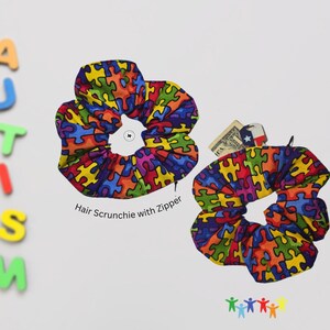 May include: Two colorful hair scrunchies with a puzzle piece pattern. The scrunchies are made of fabric and have a zipper. The text "Hair Scrunchie with Zipper" is printed below the scrunchies.