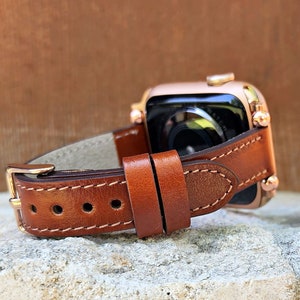 May include: A brown leather watch band with a gold buckle is attached to a rose gold smartwatch. The band has white stitching and is set against a neutral background. The watch face is visible.