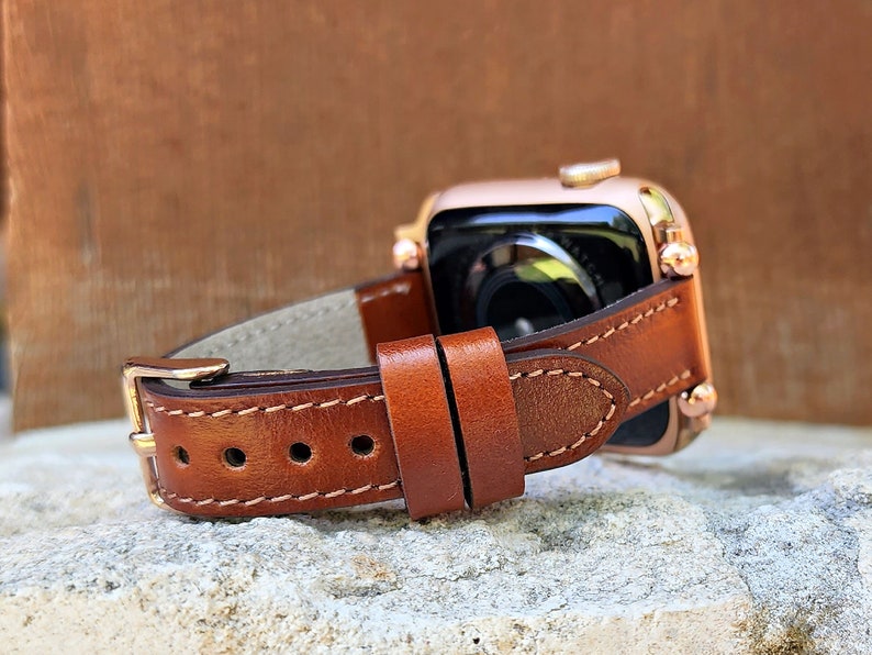 May include: A brown leather watch band with a gold buckle is attached to a rose gold smartwatch. The band has white stitching and is set against a neutral background. The watch face is visible.