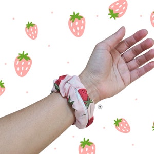 Peut inclure: Un chouchou rose avec un motif de fraises est enroul&eacute; autour d'un poignet. Le chouchou a une fermeture &eacute;clair. Le fond est blanc avec des illustrations de fraises roses.