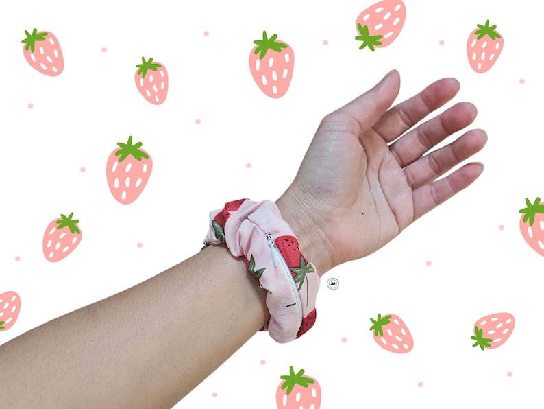 Peut inclure: Un chouchou rose avec un motif de fraises est enroul&eacute; autour d'un poignet. Le chouchou a une fermeture &eacute;clair. Le fond est blanc avec des illustrations de fraises roses.