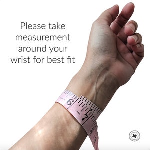 May include: A pink measuring tape wrapped around a person's wrist. The tape reads "6" and "7" inches. The text "Please take measurement around your wrist for best fit" is visible at the top of the image.