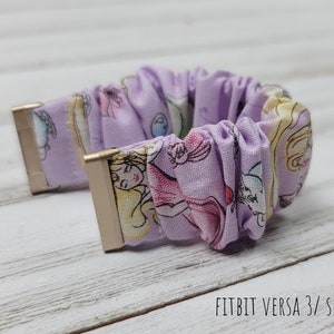 May include: Purple and white patterned watch band with gold accents. The band is made of a soft, stretchy fabric and is designed for the Fitbit Versa 3/Sense.  FITBIT VERSA 3/ SENSE text is visible on the bottom of the image.