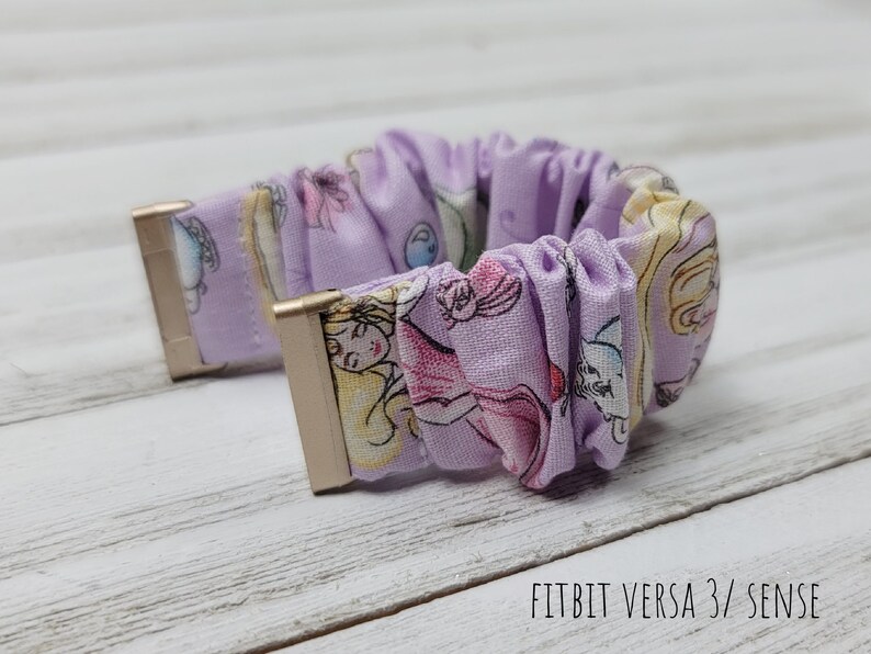 May include: Purple and white patterned watch band with gold accents. The band is made of a soft, stretchy fabric and is designed for the Fitbit Versa 3/Sense.  FITBIT VERSA 3/ SENSE text is visible on the bottom of the image.