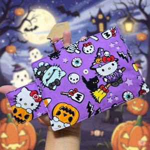 May include: A purple Halloween-themed wallet featuring Hello Kitty characters in witch costumes and other spooky elements. The wallet has a matching card holder and is set against a Halloween night background with pumpkins and a full moon.