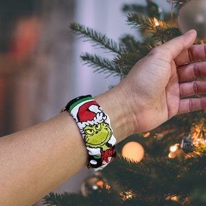 May include: A white and green watch strap with a cartoon image of the Grinch wearing a Santa hat. The strap is on a person's wrist.