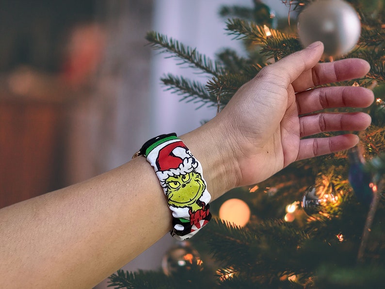 May include: A white and green watch strap with a cartoon image of the Grinch wearing a Santa hat. The strap is on a person's wrist.