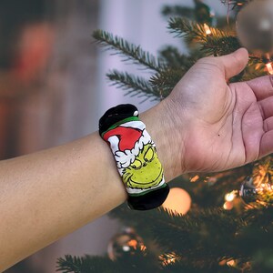 May include: A black and green wristband featuring a cartoon image of the Grinch wearing a Santa hat. The wristband is worn on a person's wrist.