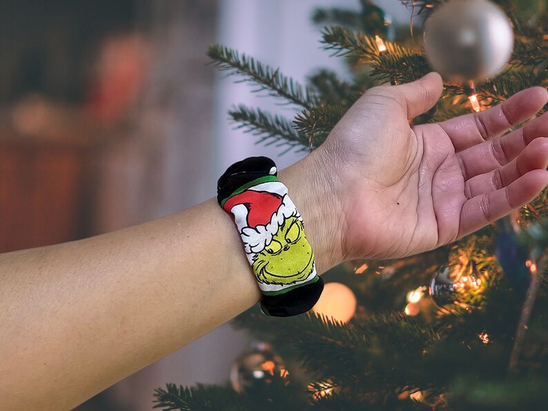 May include: A black and green wristband featuring a cartoon image of the Grinch wearing a Santa hat. The wristband is worn on a person's wrist.