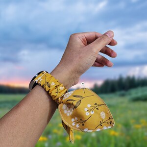 Puede incluir: Una correa de reloj de tela color amarillo mostaza con un diseño elástico tipo coletero y un lazo decorativo. La tela tiene un estampado de racimos florales blancos y tallos negros finos con hojas, unida a un conector de reloj metálico.