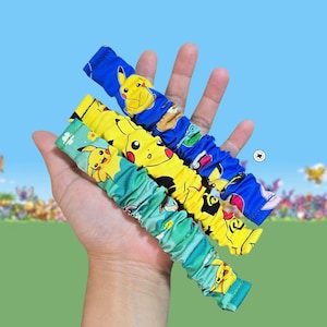 May include: Three fabric scrunchies featuring Pikachu character prints. The top scrunchie is blue, the middle is yellow, and the bottom is teal green. Each scrunchie has a gathered, elasticized design with repeating yellow Pikachu illustrations.