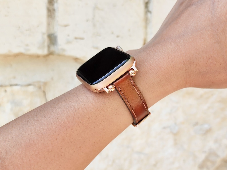 May include: A rose gold smartwatch with a brown leather band is displayed on a wrist. The watch face is black. The band is stitched and has a buckle. The watch case has decorative gold accents.