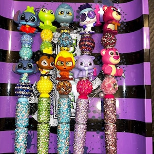 May include: A collection of decorative pens featuring character toppers. Each pen has a jeweled barrel in various colors, topped with a character figurine and a decorative bead. The pens are arranged on a purple tray with black stripes.