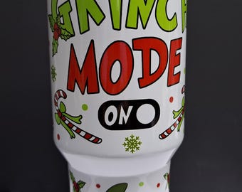Puede incluir: Un vaso de acero inoxidable blanco con tapa transparente, con un diseño de goteo de baba verde en la parte superior. El texto dice GRINCH MODE ON en letras verdes y rojas. La sección inferior muestra un personaje de dibujos con gorro de Navidad.