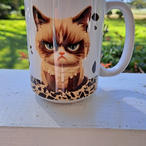 Op de afbeelding: Witte keramische koffiemok met een illustratie van een chagrijnige kat. De kat heeft bruine vacht, groene ogen en een zwarte neus. De mok heeft een zwart handvat en een bruin en wit gepatroneerde bodem.