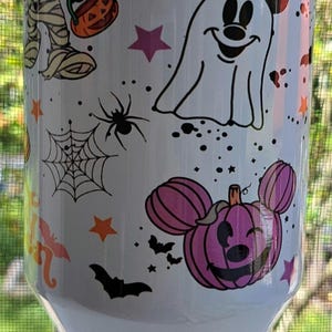 Puede incluir: Vaso blanco con un dise&ntilde;o de Halloween con personajes de dibujos animados. El dise&ntilde;o incluye una momia, un fantasma, telara&ntilde;as, murci&eacute;lagos, estrellas y calabazas con forma de Mickey Mouse. El vaso est&aacute; decorado con detalles en negro, naranja, morado y rosa.
