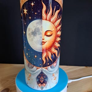 Puede incluir: Un vaso de acero inoxidable con un diseño colorido que presenta un sol y una luna con un fondo de noche estrellada. El vaso está en un soporte giratorio blanco y azul.