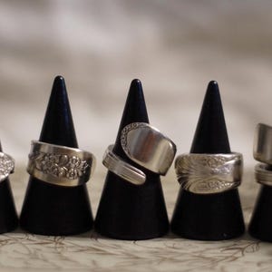 May include: Five silver rings made from recycled silverware displayed on black stands. The rings are all different designs, some with floral patterns and some with plain bands.