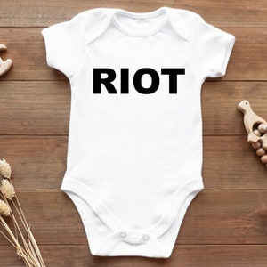May include: White baby onesie with black text that reads "RIOT". The onesie is on a wooden surface with a wooden toy horse and a wooden toy bird.